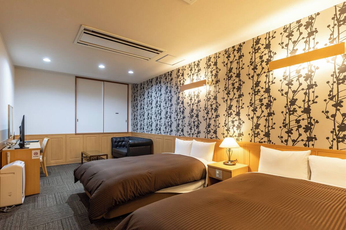 A hotel room with two beds, a desk, a chair, a wardrobe, and a floral wallpaper.