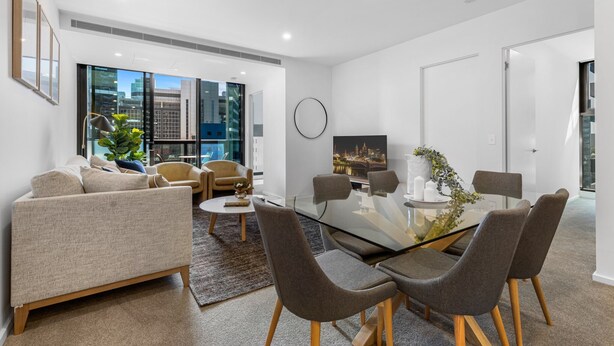 Un moderno salón con una mesa de comedor de vidrio, sillas tapizadas en gris, un sofá color beige y un amplio ventanal con vistas a la ciudad.