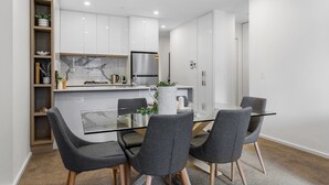 Una cocina moderna con armarios blancos, un refrigerador de acero inoxidable y una mesa de comedor de vidrio con cuatro sillas grises.