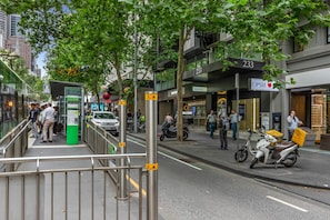 Eine belebte Stadtstraße mit Fußgängern, einem Lieferdienst auf einem Roller und Geschäften, darunter ein Geschäft namens 'SNCOP' und 'OPS'.