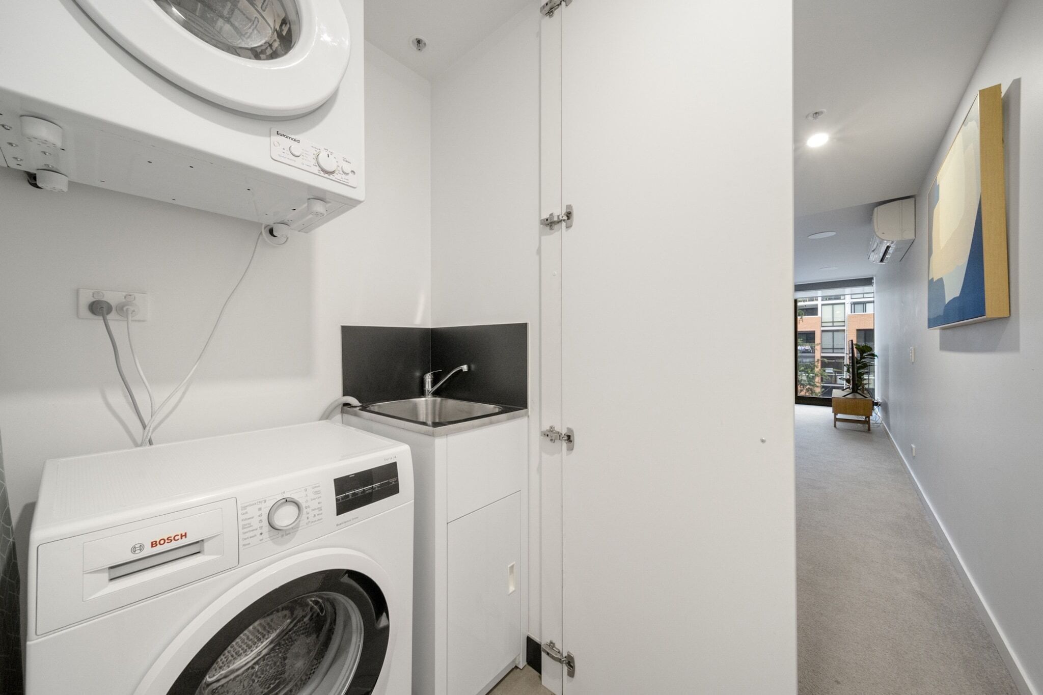 Een praktische wasruimte met een Bosch wasmachine en droger, een wastafel en een deur die uitkomt op een gang met uitzicht op gebouwen.