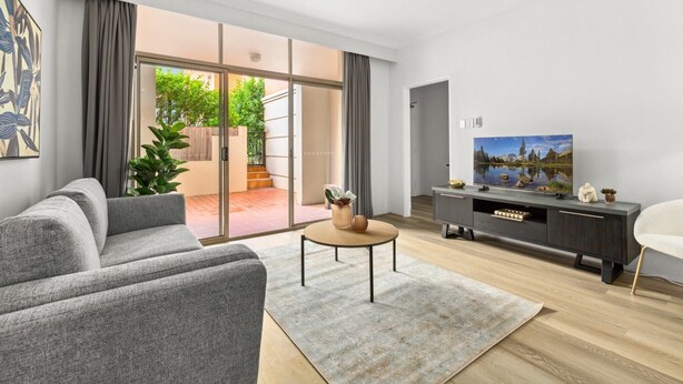 A modern living room with a grey sofa, a coffee table, a TV on a dark wooden cabinet, and a sliding glass door leading to an outdoor area with greenery. - Bright Inner-West Stay with Pool Access (Camperdown)