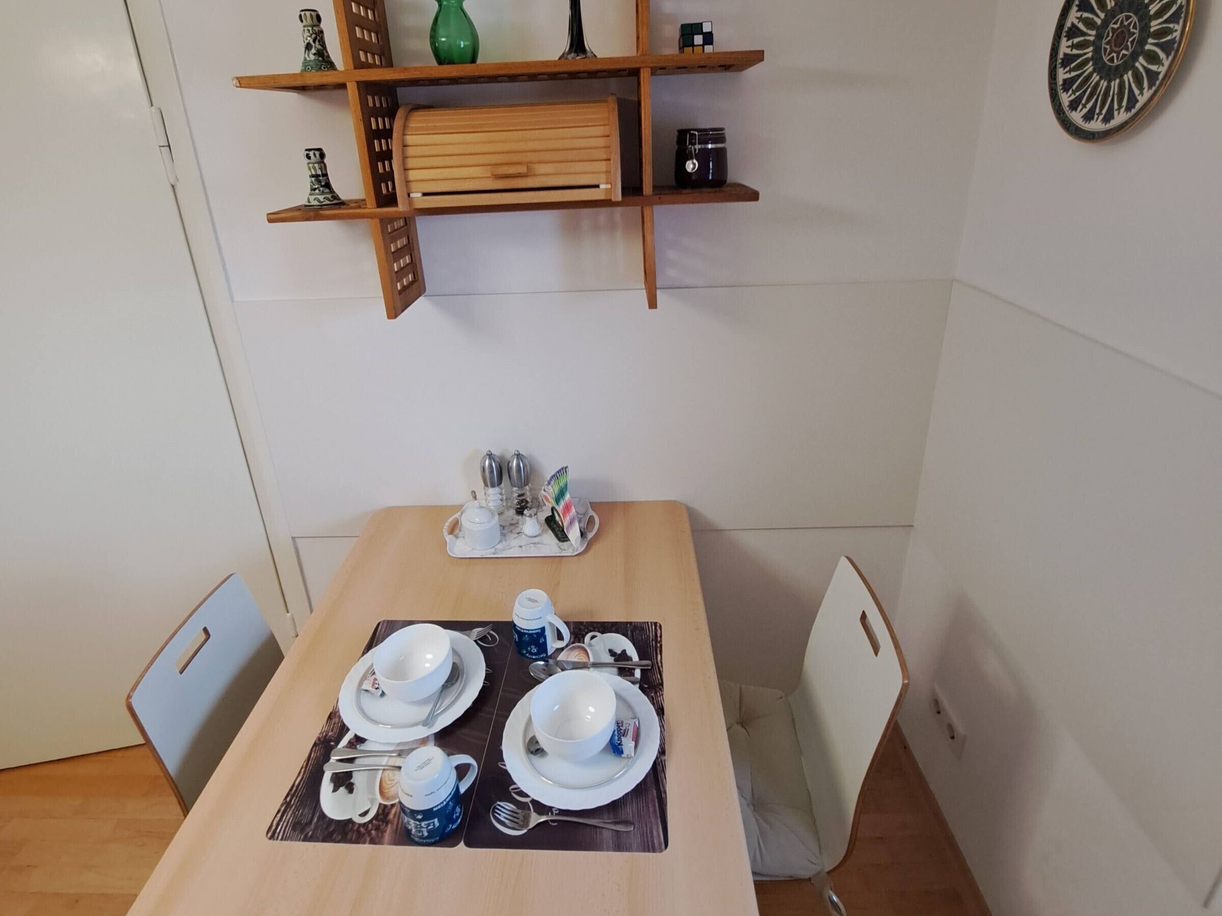 A dining area with a table set for two, including plates, cups, and cutlery. There is a shelf above the table with decorative items and a small clock.