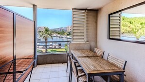 A balcony with a wooden table and chairs, a metal railing, and a view of a marina and buildings. - Waterfront Riviera Charm with a View (Mandelieu-la-Napoule)
