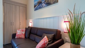 A modern living room with a dark leather sofa, red patterned cushions, a potted plant, and a painting of a beach scene on the wall.