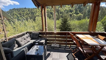 A covered outdoor seating area with a sofa, table, and chairs, overlooking a forested landscape.