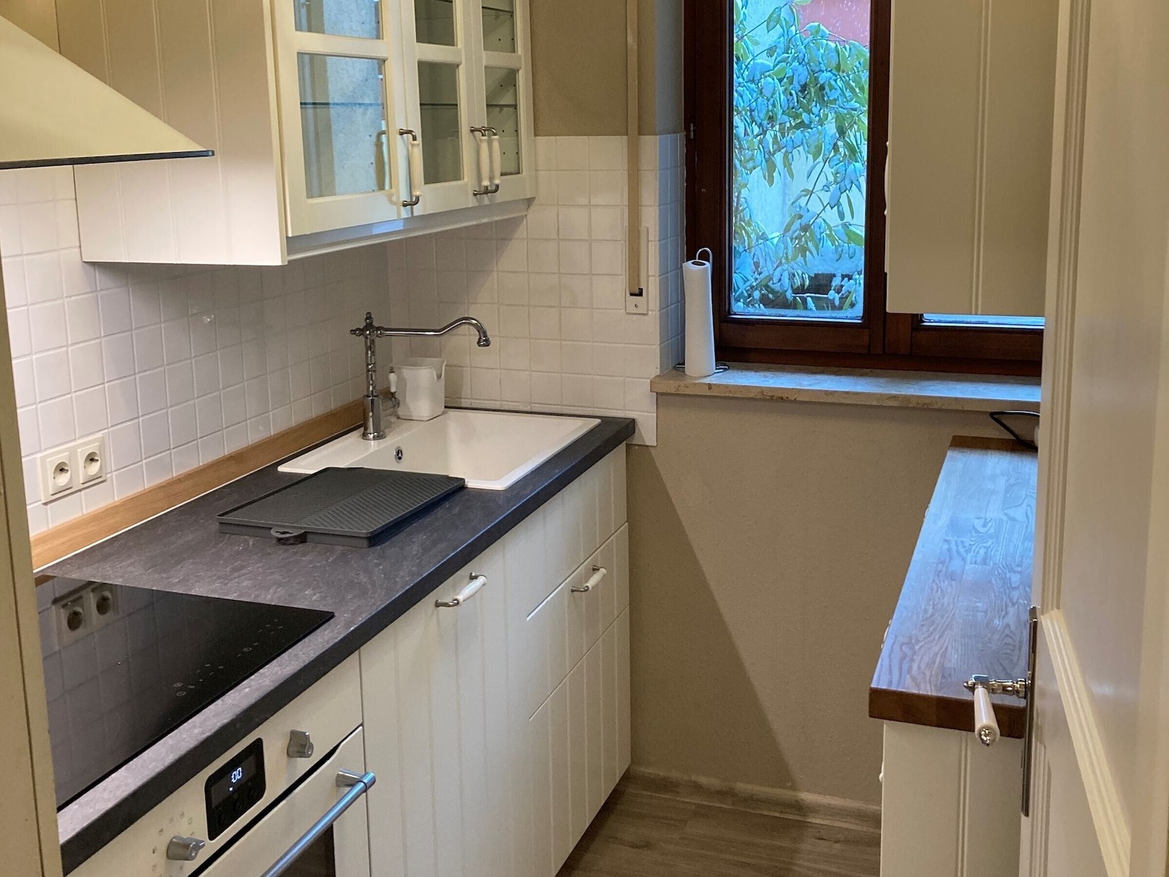 Eine kleine Küche mit weißen Schränken, einer schwarzen Arbeitsplatte, einem Fenster mit Blick auf Grünflächen und einem eingebauten Backofen.