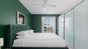 Un dormitorio con una cama tapizada de verde, ropa de cama blanca, un ventilador de techo, una obra de arte enmarcada y vistas a través de una puerta de cristal.