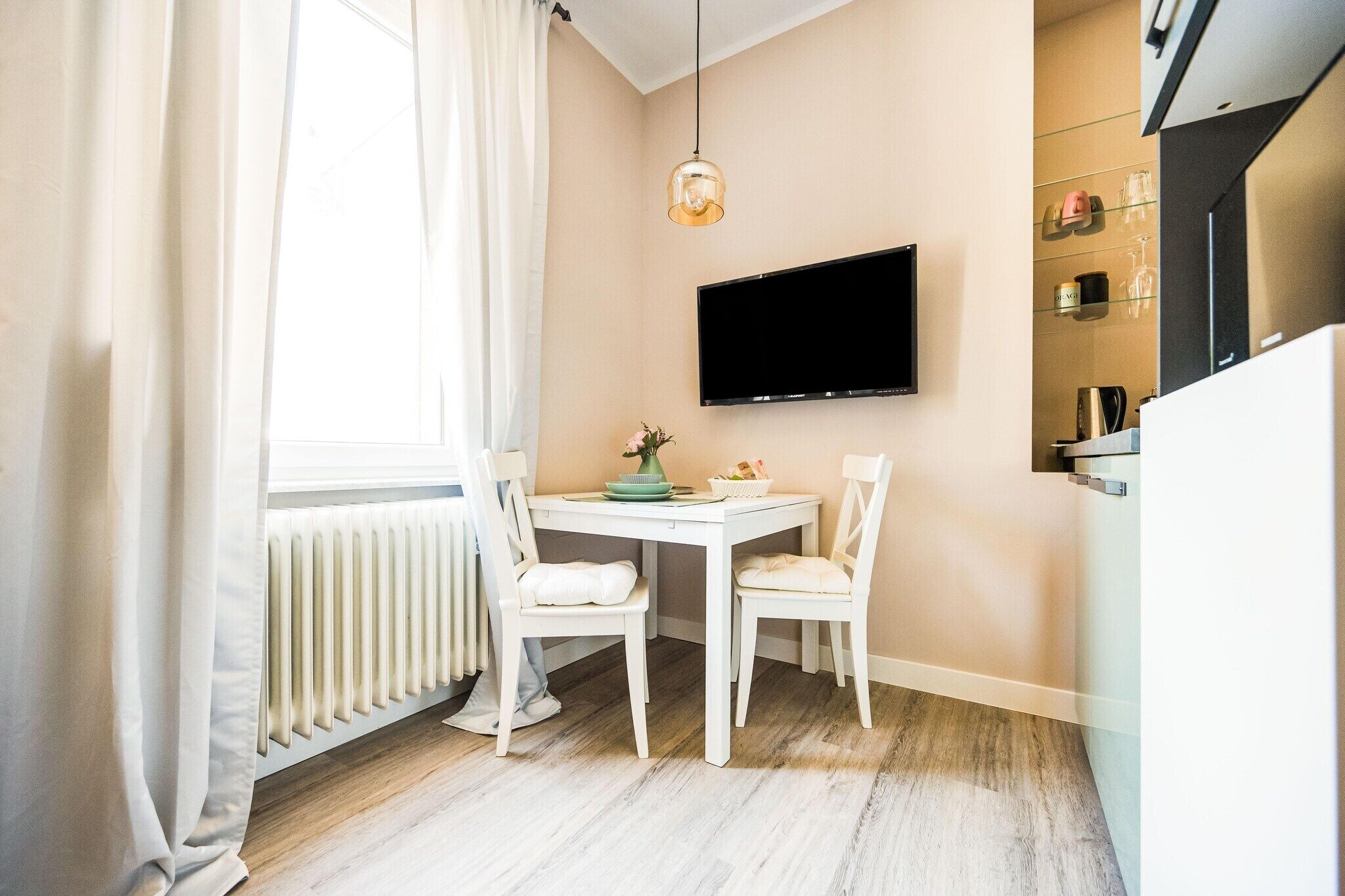 Un pequeño comedor con una mesa y sillas blancas, un televisor de pantalla plana montado en la pared y una ventana con cortinas.