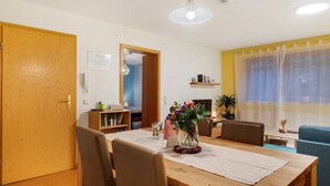 Un comedor con una mesa de madera, sillas, un florero con flores, una estantería y una ventana con cortinas.