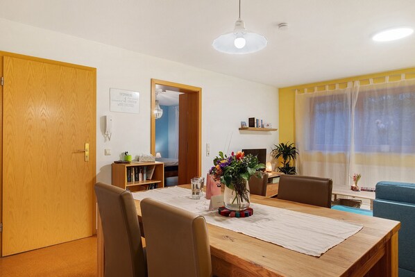 Ein Essbereich mit Holztisch, Stühlen, einer Blumenvase, einem Bücherregal und einem Fenster mit Vorhängen.