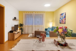 Ein Wohnzimmer mit Fernseher, Couch, einem Couchtisch mit Gläsern und einer Vase mit Blumen.