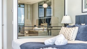 Une chambre d’hôtel moderne avec un grand lit, une cloison en verre, une vue sur la ville et une table de chevet avec une lampe.