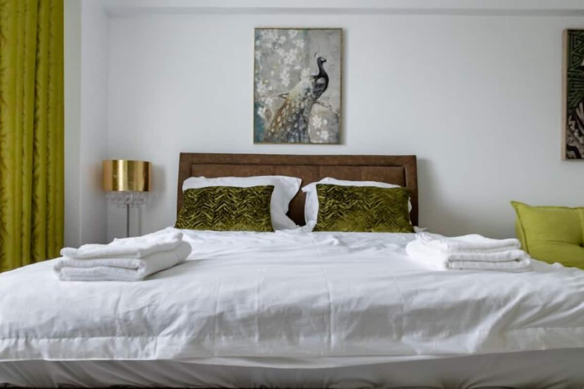 Un lit bien fait, avec du linge de lit blanc et deux coussins à motifs verts, une tête de lit en bois et deux œuvres d’art accrochées aux murs.