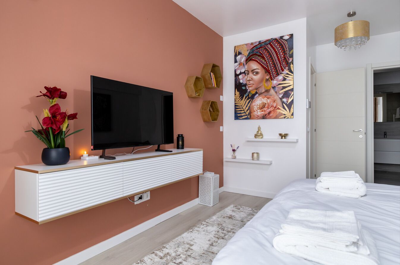 Une chambre d’hôtel moderne avec un grand lit, un téléviseur à écran plat, un tableau au mur et une console blanche sur laquelle est posé un vase de fleurs.