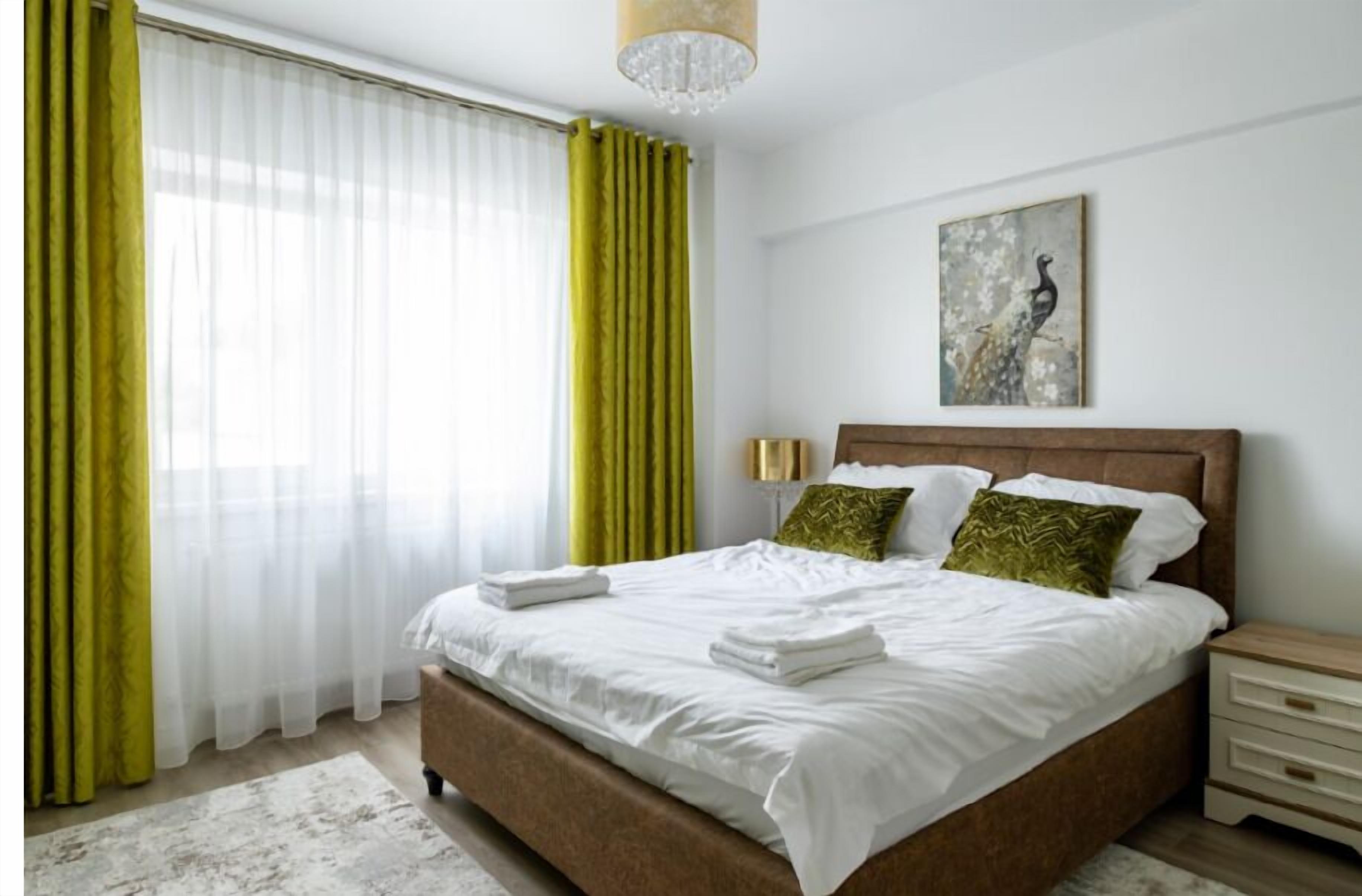 Ein ordentlich bezogenes Bett mit weißen Bettwäsche, einem Holz-Kopfteil und zwei Kissen. Ein Nachttisch mit Lampe und ein kleiner Nachttisch mit Schubladen sind vorhanden. Ein Vogelbild hängt an der Wand über dem Bett.