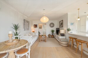 A modern living room with a wooden dining table, white sofa, and a TV. There are plants, a lamp, and a hanging light fixture. - Sunrise Cove 2-Bed Apartment at Newport Beach (Newport)