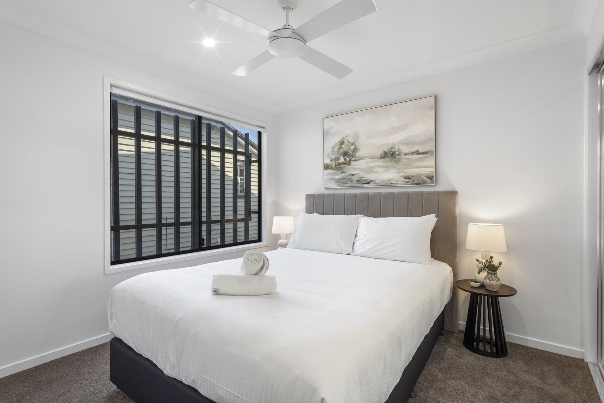A bedroom with a bed, a ceiling fan, a window with blinds, a painting on the wall, and a bedside table with a lamp.