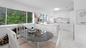 Una cocina moderna con armarios blancos, una mesa de comedor de vidrio y vistas a la vegetación a través de las ventanas.