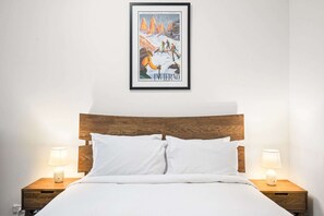 A neatly made bed with white linens and two bedside lamps on wooden nightstands. Above the bed, there is a framed poster with a mountainous landscape and the word "INVERNO". - Peaceful 2-Bed Apartment with Gym & Pool (West Melbourne)