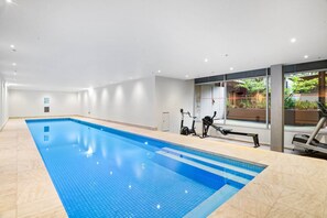 A swimming pool with exercise equipment, a view of greenery, and a modern interior design. - Peaceful 2-Bed Apartment with Gym & Pool (West Melbourne)
