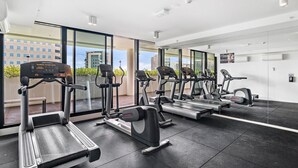A modern gym with various cardio machines, large windows, and a view of buildings outside. - South Melbourne Apartment with Gym Access (South Melbourne)