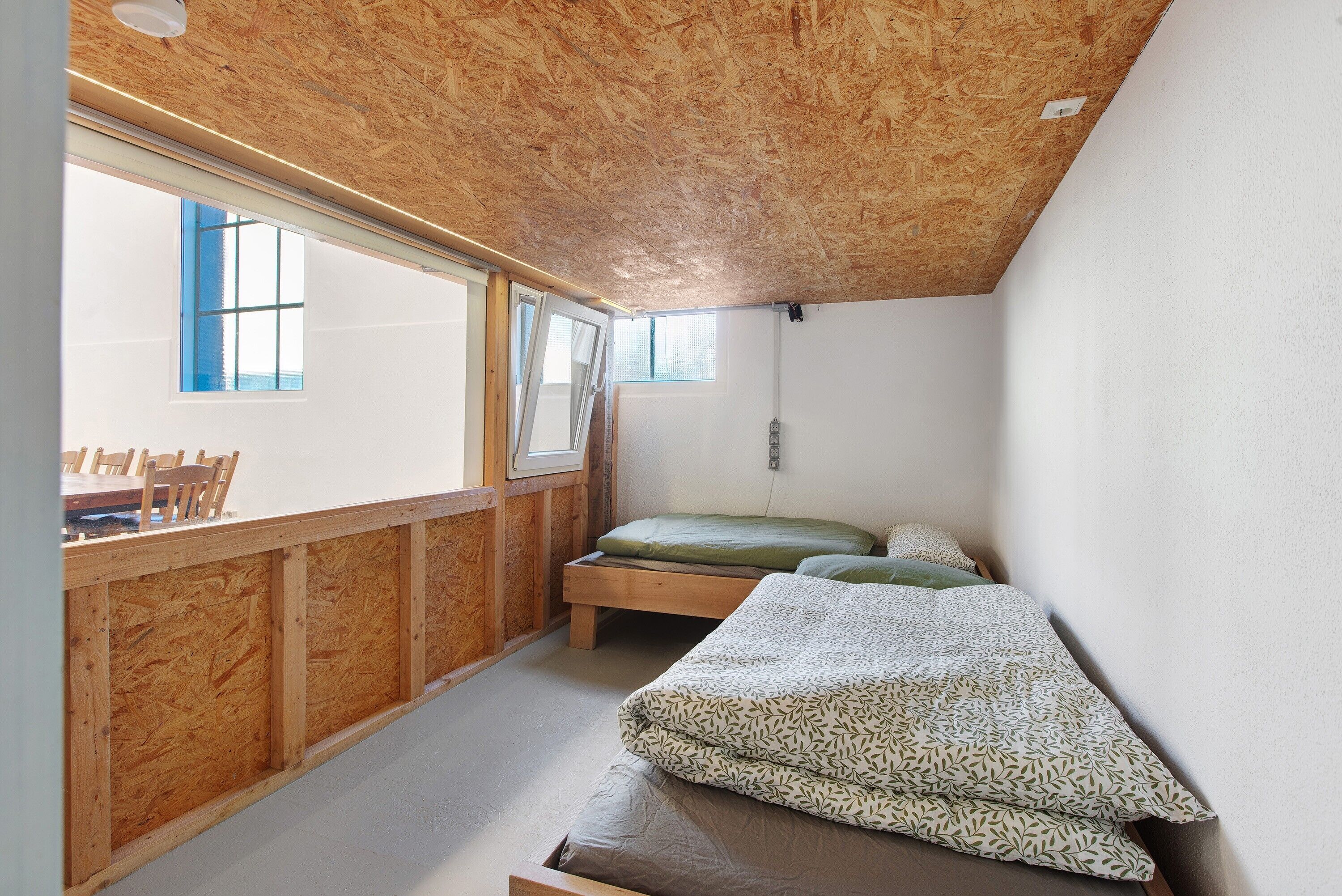 Ein kleiner, gemütlicher Raum mit Holzbalkendecke, einem Bett mit gemustertem Bettzeug, einem Fenster mit blauen Rollläden und einer hölzernen Sitzbank.