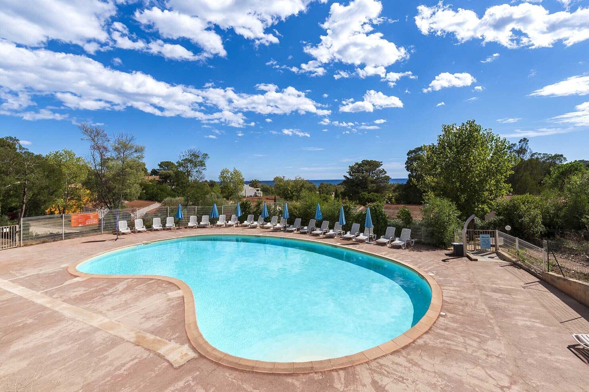 A swimming pool with lounge chairs and umbrellas, surrounded by trees and a clear sky.