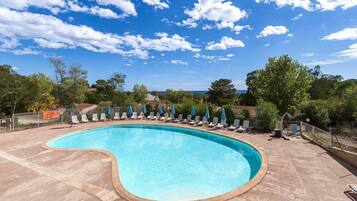 A swimming pool with lounge chairs and umbrellas, surrounded by trees and a clear sky.