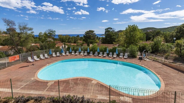 A large, curved swimming pool with lounge chairs and umbrellas, surrounded by a fence, with a view of the sea and hills in the background. - Coastal and Garden Living in Corsica (Sari-Solenzara)
