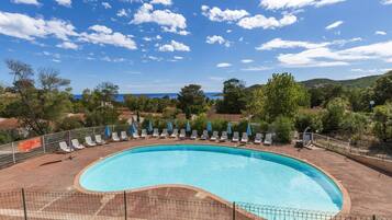 A large, curved swimming pool with lounge chairs and umbrellas, surrounded by a fence, with a view of the sea and hills in the background.