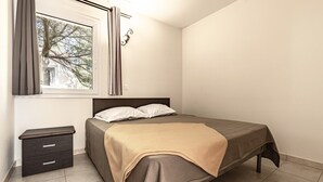 A bedroom with a bed, a nightstand, a window with curtains, and a view of trees outside. - Coastal and Garden Living in Corsica (Sari-Solenzara)