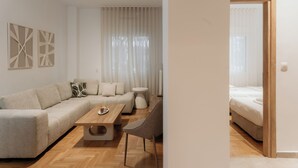 Una sala de estar moderna con un sofá, una mesa de centro y una silla. Se ve una cama al fondo, junto a una ventana con cortinas.