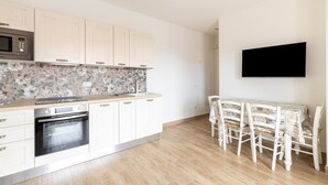 Une cuisine avec des meubles blancs, un four à micro-ondes, un four traditionnel, une table à manger avec des chaises et un téléviseur à écran plat fixé au mur.