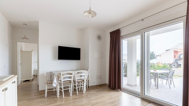 Une pièce spacieuse avec un plancher en bois, une table à manger avec des chaises, un téléviseur à écran plat fixé au mur et une porte-fenêtre coulissante donnant sur un espace extérieur.