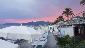 Eine Seeterrasse mit Reihen aus weißen Liegestühlen und Sonnenschirmen, mit Blick auf eine gebirgige Landschaft und das Meer.