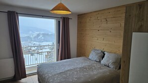 Une chambre à coucher avec un lit, une tête de lit en bois, une vue sur un paysage enneigé depuis un balcon et un luminaire suspendu.