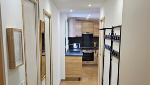 Un couloir étroit, doté d’un plancher en bois, mène à une cuisine avec des meubles de rangement en bois et un comptoir noir.