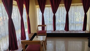 A room with a wooden table and chairs, large windows with red curtains, and a view of trees outside. - D'BONN INN (Shillong)