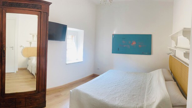 Une chambre avec une armoire en bois, un lit recouvert d’une courtepointe blanche, un téléviseur fixé au mur et une fenêtre laissant entrer la lumière naturelle.