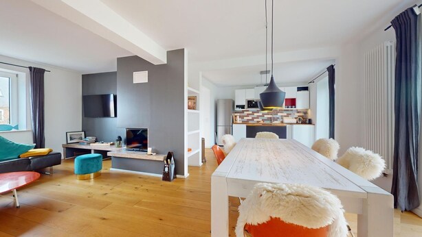Uma sala de estar moderna com piso de madeira, um sofá cinza, um banquinho vermelho, uma mesa de jantar branca com almofadas brancas fofas e uma cozinha ao fundo.