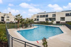 A rectangular swimming pool with a surrounding fence, palm trees, and a building with balconies in the background. - The Kings Residence: Business Ready alquiler temporal (San Sebastián de los Reyes)