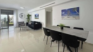 A modern living room with a dining area, featuring a large window, a sofa, a coffee table, and a dining table with chairs. - Ariston  First Line Lake View (Tiberias)