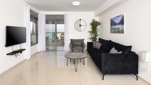 A modern living room with a flat-screen TV, a black sofa, a grey armchair, a small round coffee table, and a potted plant. - Ariston  First Line Lake View (Tiberias)