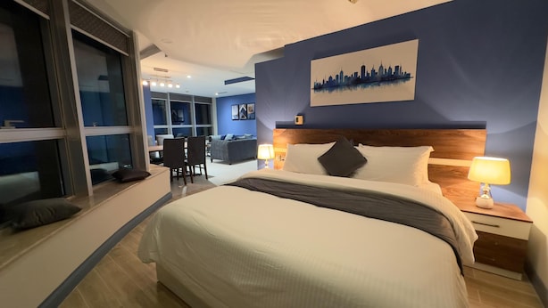 A modern hotel room with a large bed, a city skyline painting on the wall, and a dining area in the background. - The Grand Skyline Corner Suite (Islamabad)