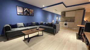 A modern living room with a grey sectional sofa, a wooden coffee table, and a kitchen area with a washing machine. - The Grand Skyline Corner Suite (Islamabad)