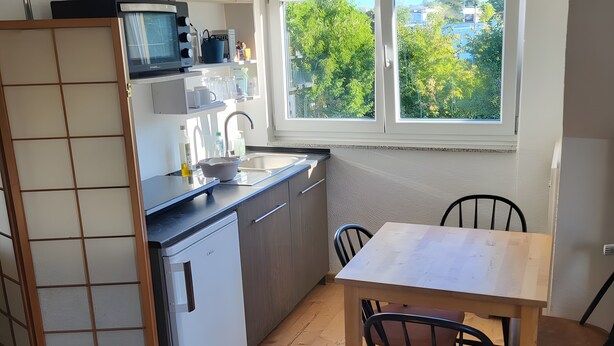 Cocina compacta con microondas, lavamanos y armarios, una mesa de comedor con sillas y una ventana con vistas a árboles.