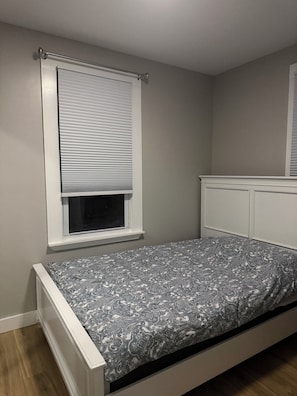 Une chambre à coucher avec un lit, une fenêtre avec des stores, une tête de lit blanche et un radiateur fixé au mur.