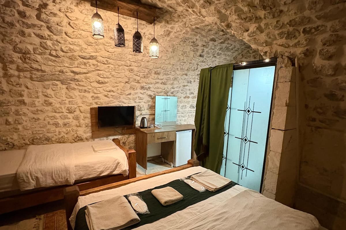 Ein Zimmer mit Steinwand, zwei Betten, einem kleinen Schreibtisch, einem Spiegel und einem fenster mit Vorhängen.