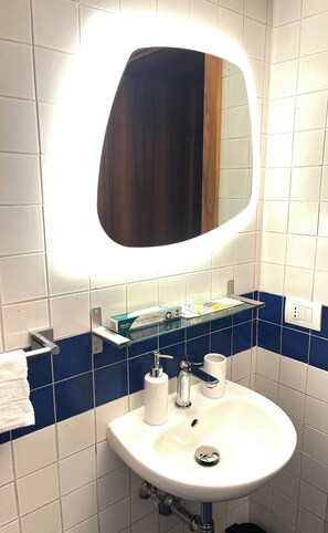 A bathroom with a white sink, a mirror with a light, a blue-tiled wall, and a shelf with toiletries. - In the heart of the historic center a few steps from the Arena and Juliet's Balcony (Verona)